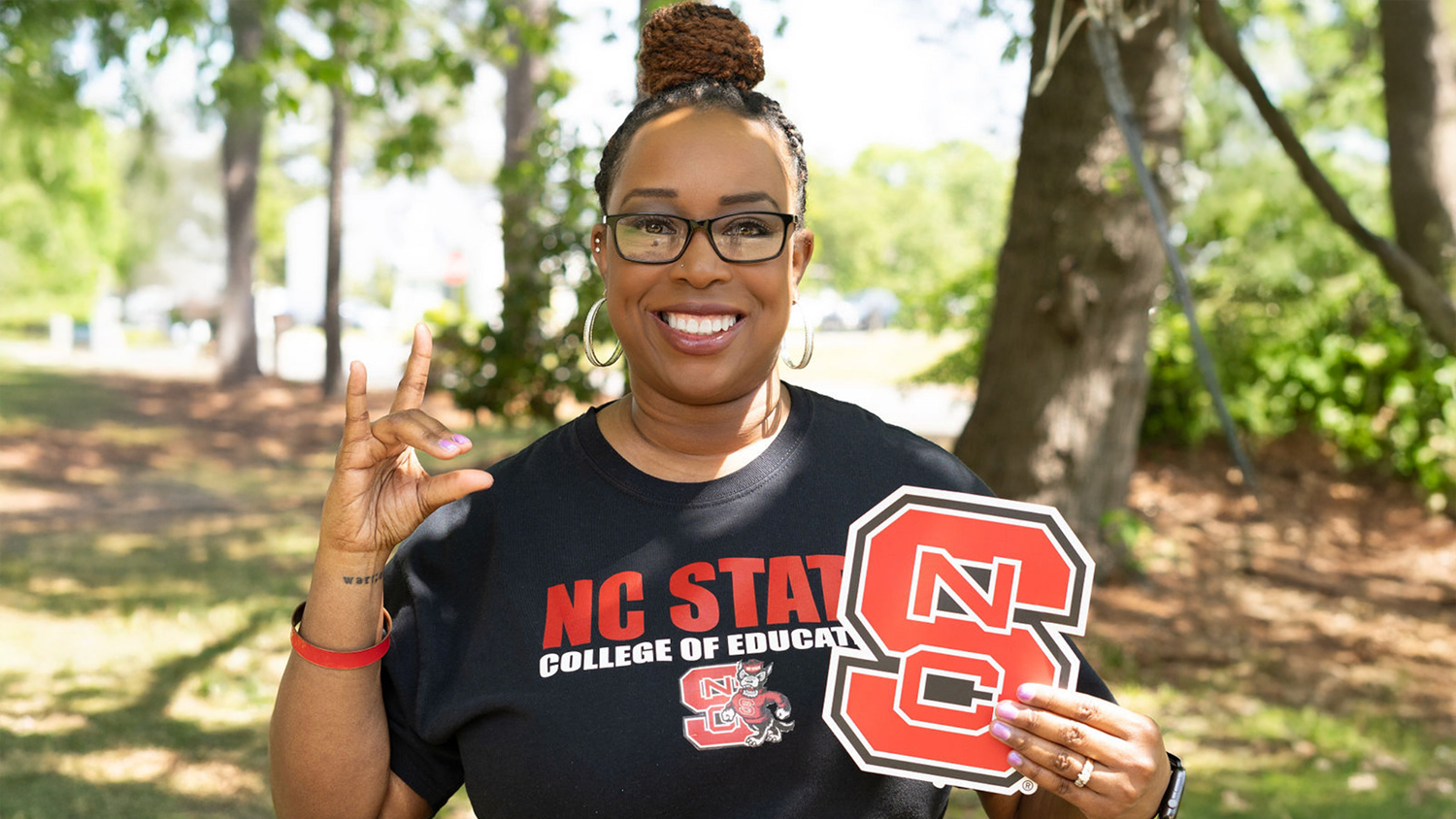 A photo of Kimberly Sutton holding up her Wolfie and a paper NC State logo while wearing an NC State College of Education T-shirt