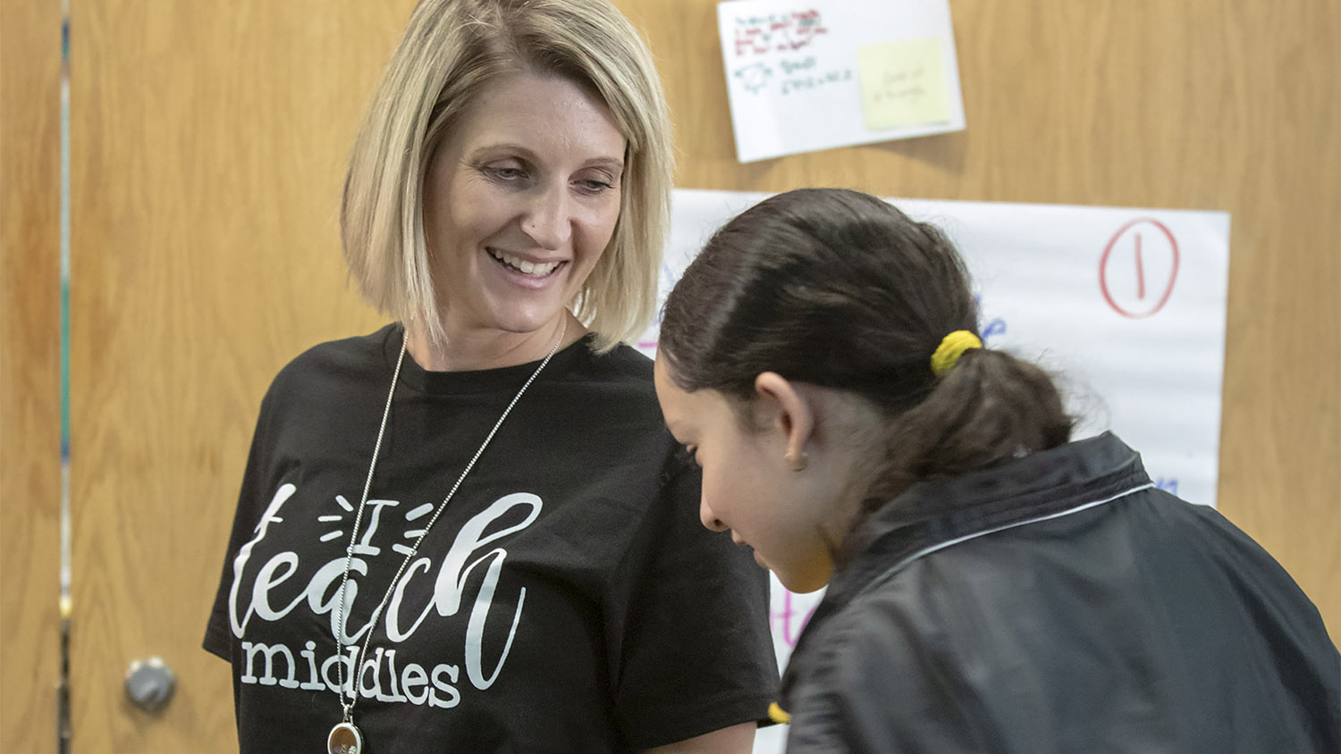 Teacher with student in a classroom. Teacher wears a black t-shirt with "I Teach Middles" messaging.