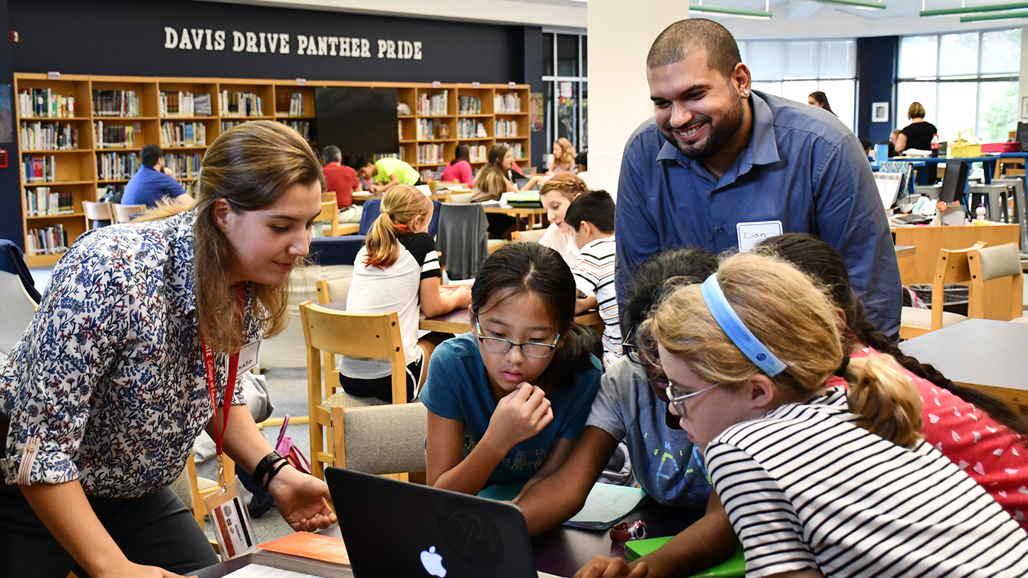 A photo of two NC State Education students mentoring a group of middle school students around a computer