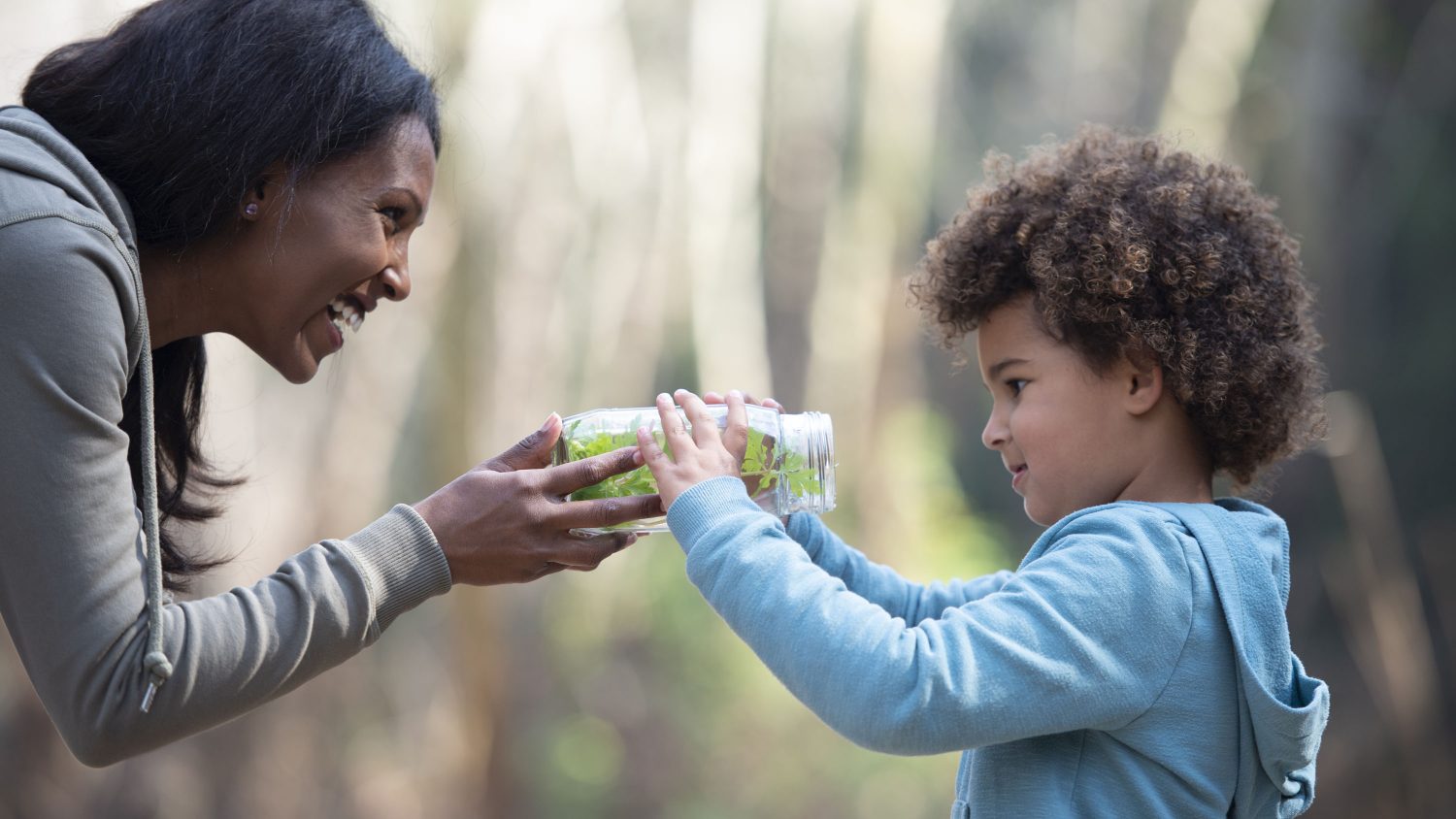 Young mother showing her 5 years old girl what inside of the jar. Mother and daughter studying nature together, holding the jar with plants inside. Teacher showing her little student jar with bugs and explaining to her about what inside. The girl looks excited.