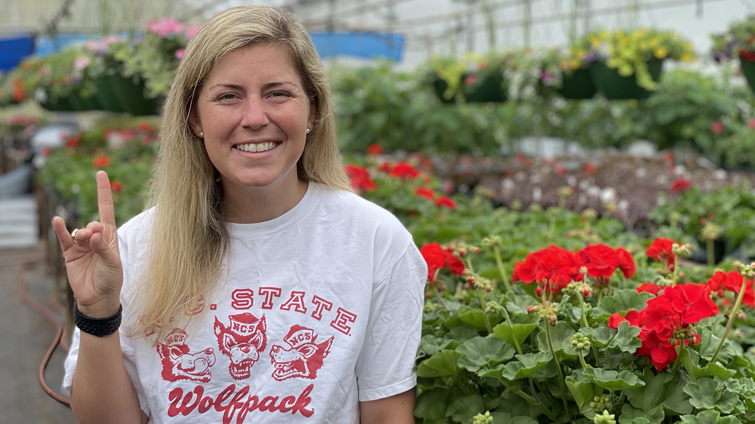 Ivy Briggs stands with the Wolfpack hand sign in a greenhouse full of flowers.