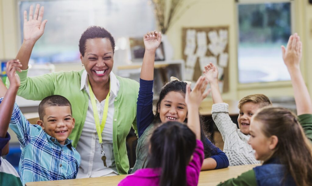 Teaching in a classroom surrounded by young students raising their hands